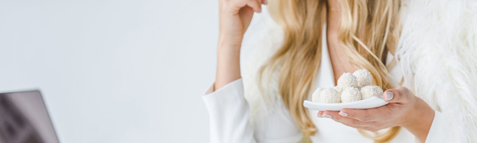 A blonde woman wearing a white shirt eats some white chocolate balls sitting in front of a laptop.