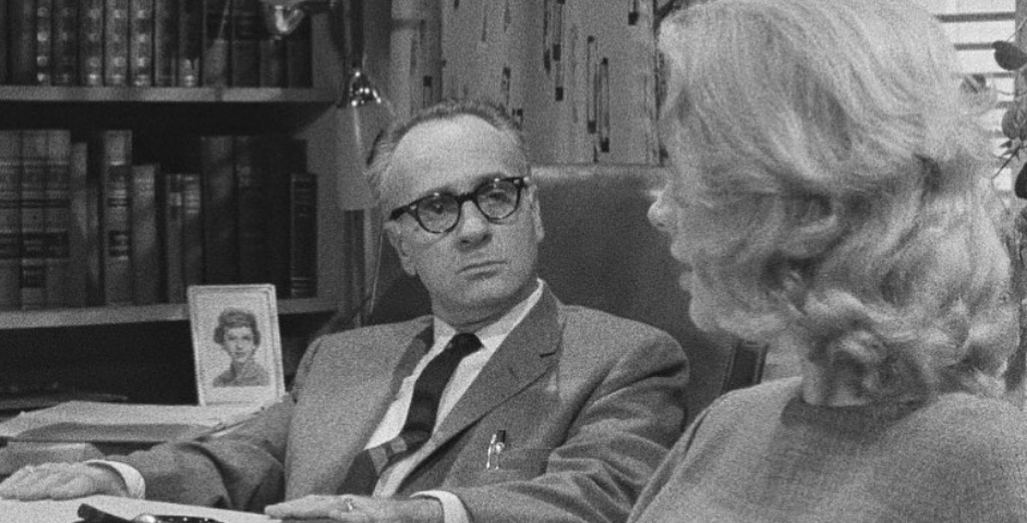 Screenshot from Carnival of Souls (1962) by  Herk Harvey. A man with glasses and a blond woman sit at a desk in a study. The subtitle reads: I have no desire for the close company of other people.