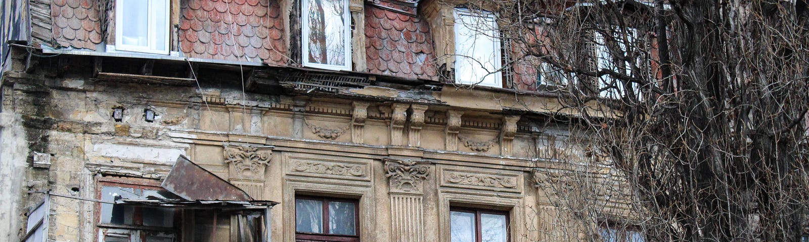 A cream-coloured stone house in obvious disrepair. There are broken roof pieces and a rusted balcony cover. A tree with no leaves is in front.