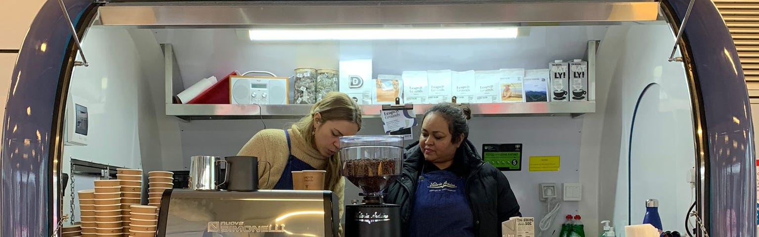 Two women are standing behind a coffee machine, inside a coffee horsebox with the name ‘Leaps & Grounds’. A menu sits on the right hand side displaying the prices.