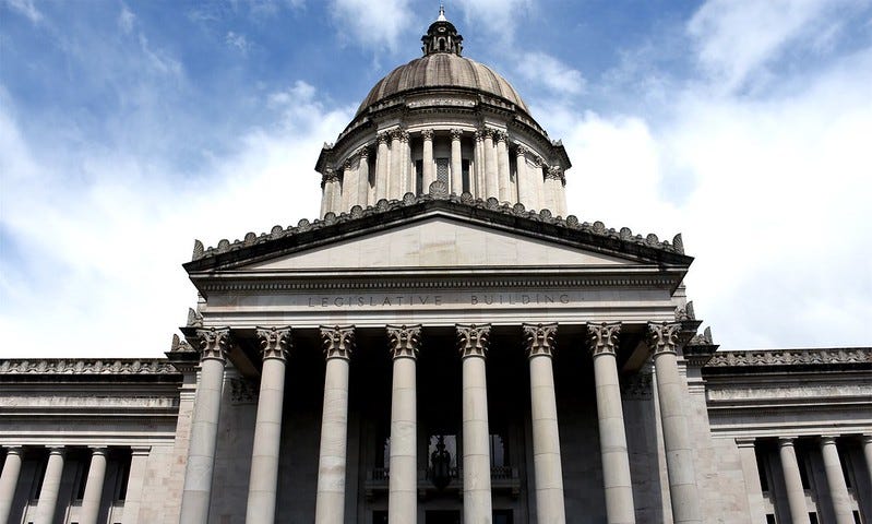 The entrance to the Washington State Legislative Building is shown. Columns adorn the entrance with the Capitol dome rising above the facade and lettering that reads, “Legislative Building.” A blue sky with clouds can be seen in the background.