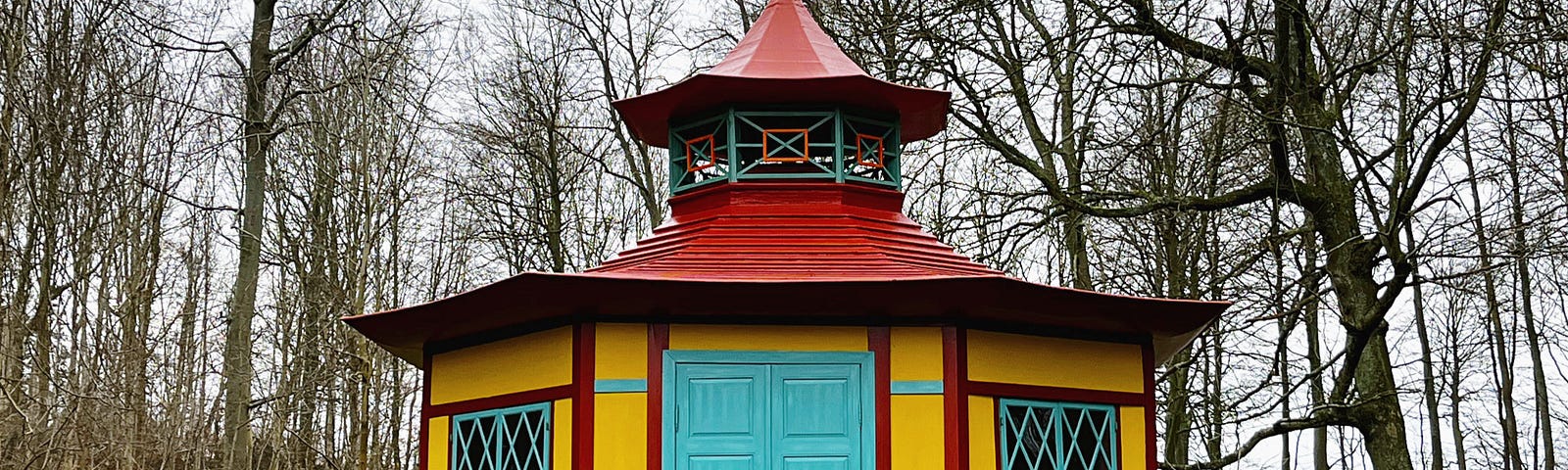 A small, hexagonal, 18th-century teahouse, painted bright yellow. with red and turquoise trim. Surrounded by bare wintery trees and set in a bright moss-green lawn.