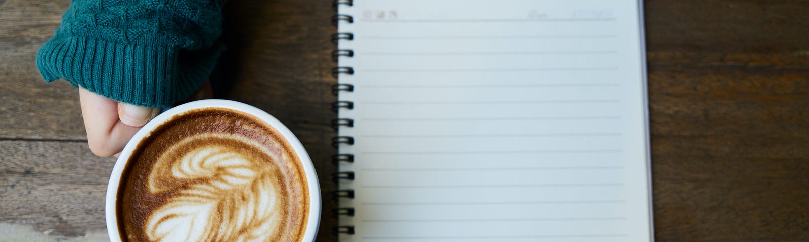 A blank notepad on a tabletop. Nearby is a hand holding a cup of coffee.