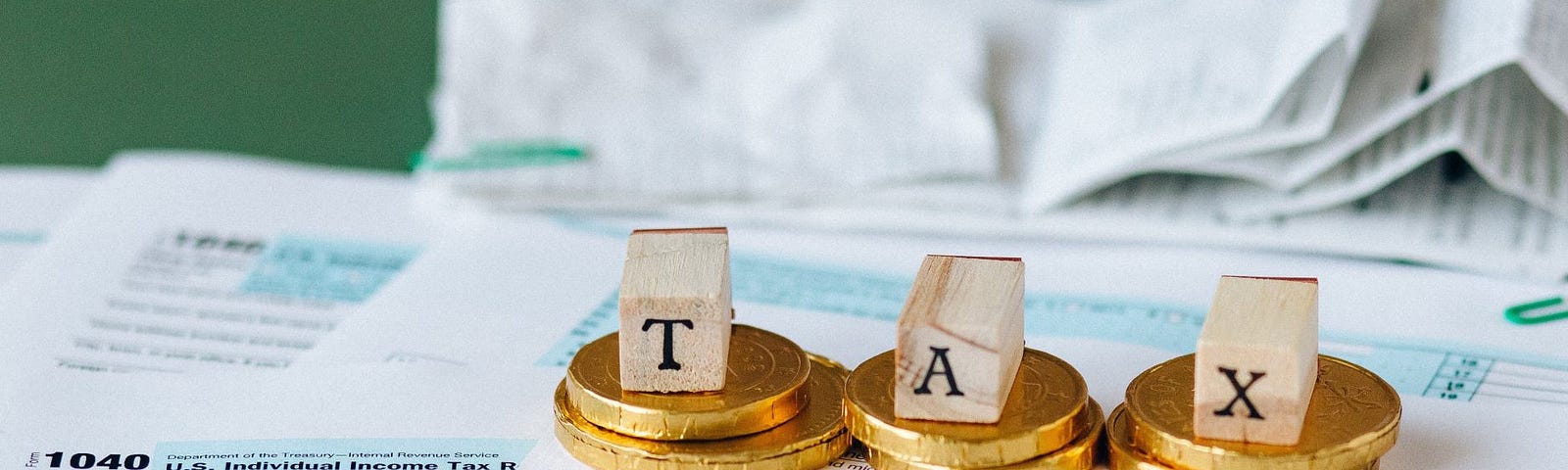 Wood blocks spelling “TAX” set atop three piles of golden coins standing on tax forms.