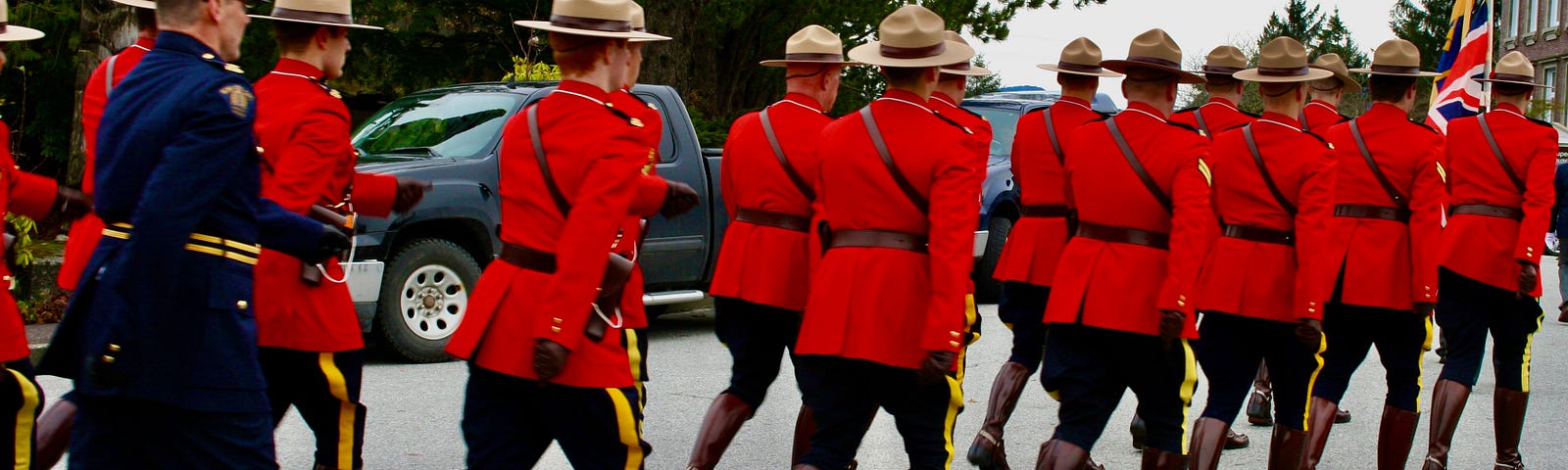 RCMP (The Royal Canadian Mounted Police) march down a street. The photo shows the backs of them walking away from the camera. They are marching in a parade.