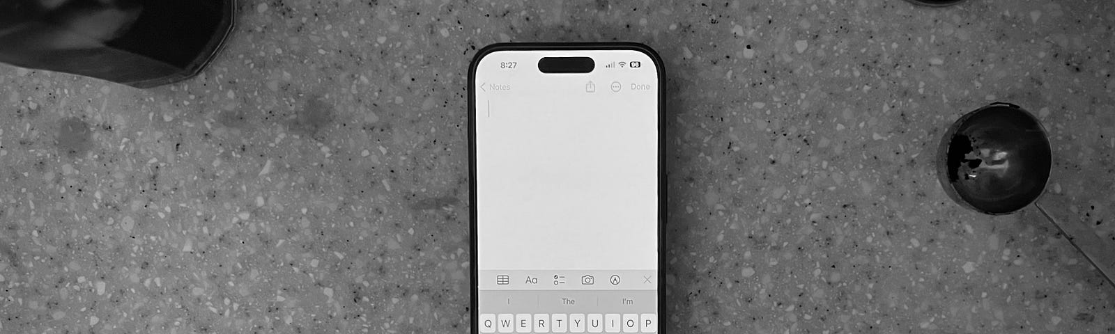 iPhone 14 Pro showing blank note with iOS Keyboard surrounded by coffee grounds and coffee tools on kitchen counter.