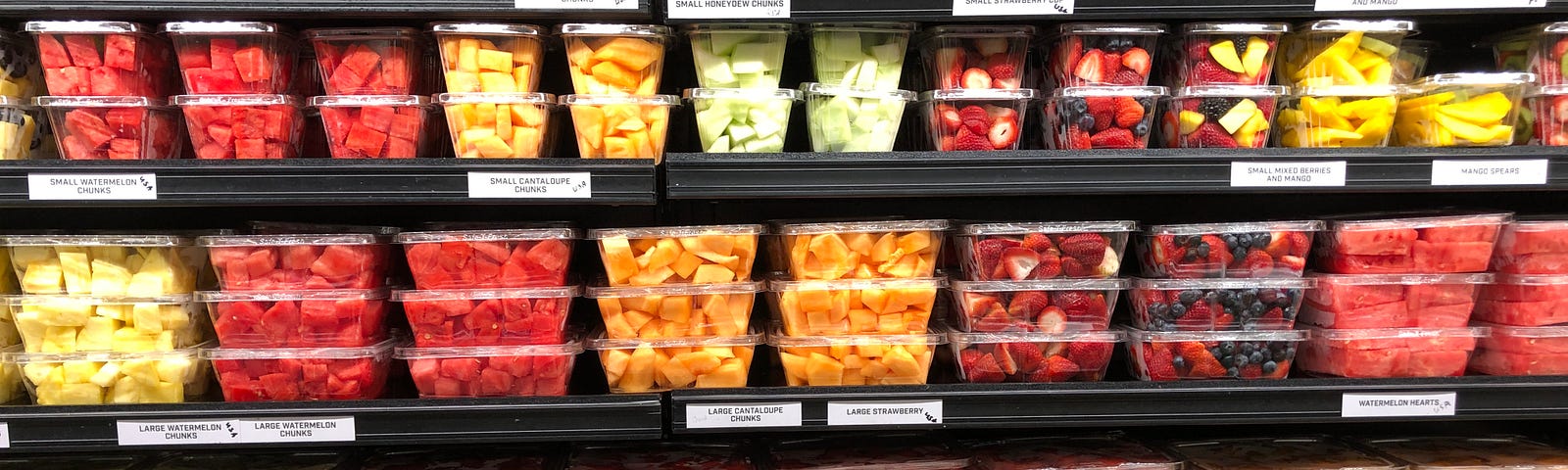 Supermarket shelves filled with overpriced plastic containers of pre-cut watermelon, pineapple, melon, and assorted fruit.