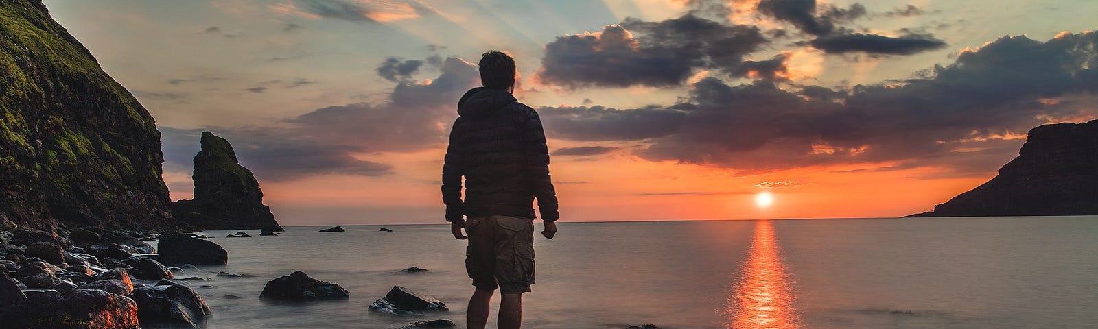 Photo of a man standing on a rock in the water. He’s wearing shorts and a long-sleeve jacket. He’s facing away from the camera and looking at a pink orange sunrise, peaking on the horizon at the distant water’s edge. The sun’s rays shine up and out, piercing the grey and white clouds in the sky. The edges of the photo show rocks and mountain in the dark foreground on the left, near the man. There’s a mountain or land in the far background on the right.