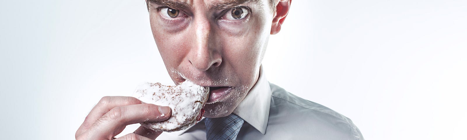 A white man wearing a white shirt and tie, taking a bit from a donut. He has a look of “I’m not sure about this” on his face, while the jelly filling has landed on his tie..