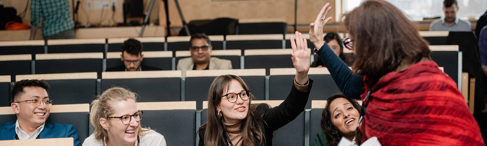 In a lecture hall, a person with a red shawl high-fives a seated person while several others look on.