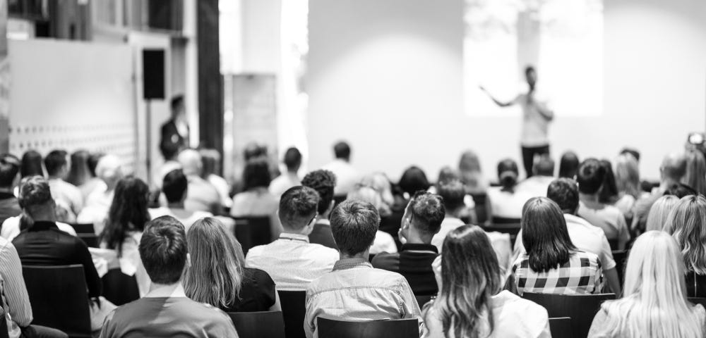 Black speaker giving a talk in conference hall to white people.