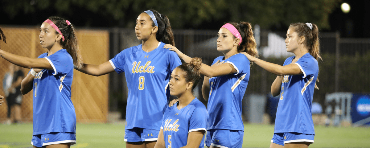 4 players on UCLA women’s soccer team toudh each other’s shoulder while 1 player of color kneels