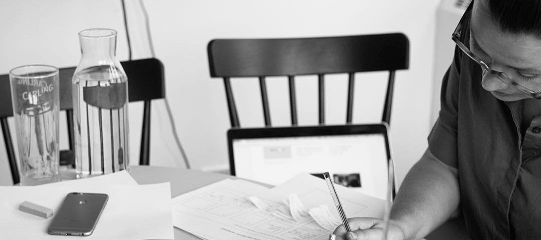 A woman sketches at a table with various bits of paper and pens around her