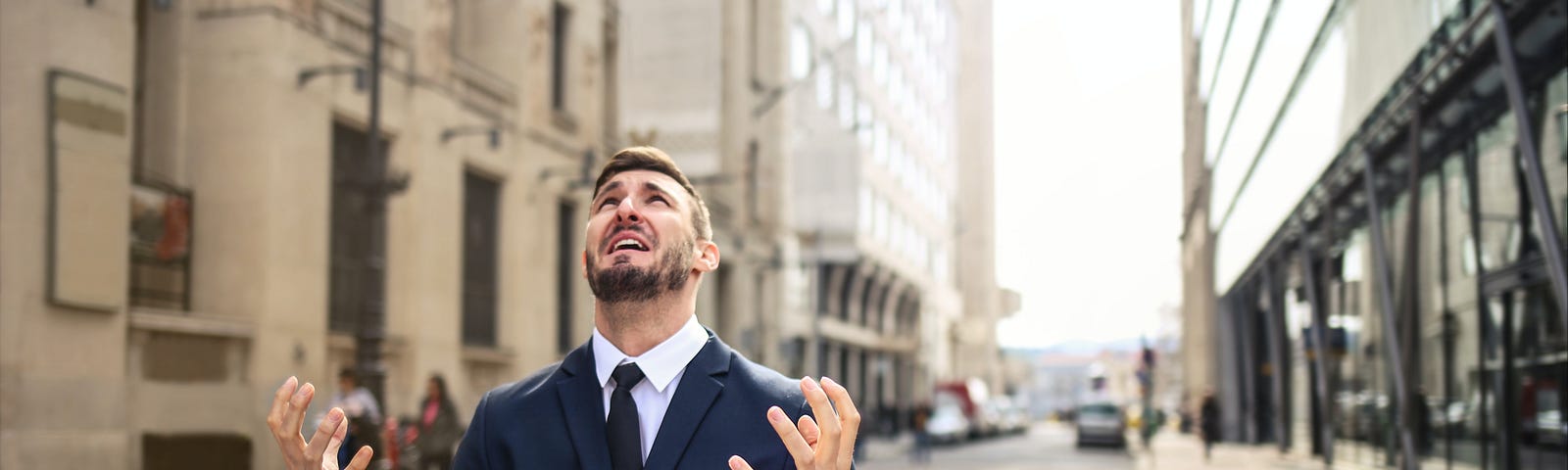 Man in suit sitting on a city street in front of a laptop clenched hands and face lifted to the sky.