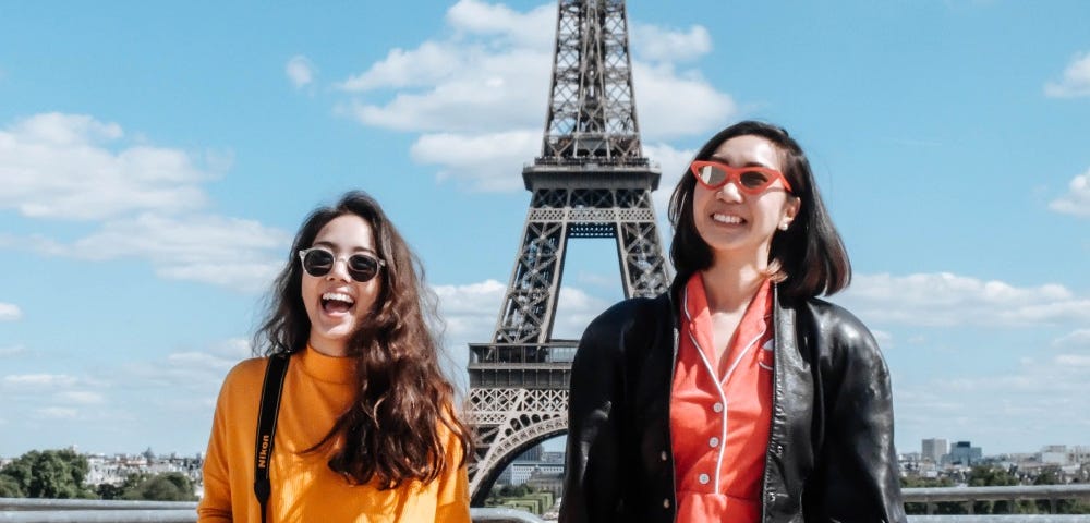 Two women smile in front of the Eiffel Tower. They wear colorful dresses and glasses.