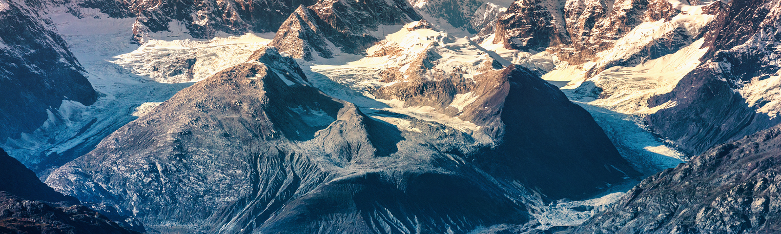 A humpback whale breaches the turquoise waters in front of a dramatic, rugged mountain range covered with glaciers and snow. The towering peaks and jagged ridges form a striking backdrop, highlighting the natural beauty of a remote and pristine Arctic landscape.