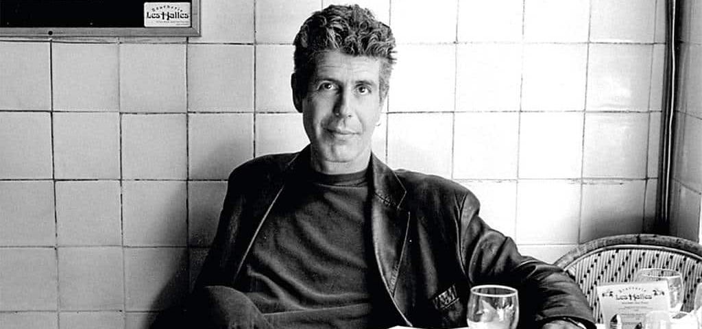 A black & white photo of Anthony Bourdain sitting at a restaurant table.