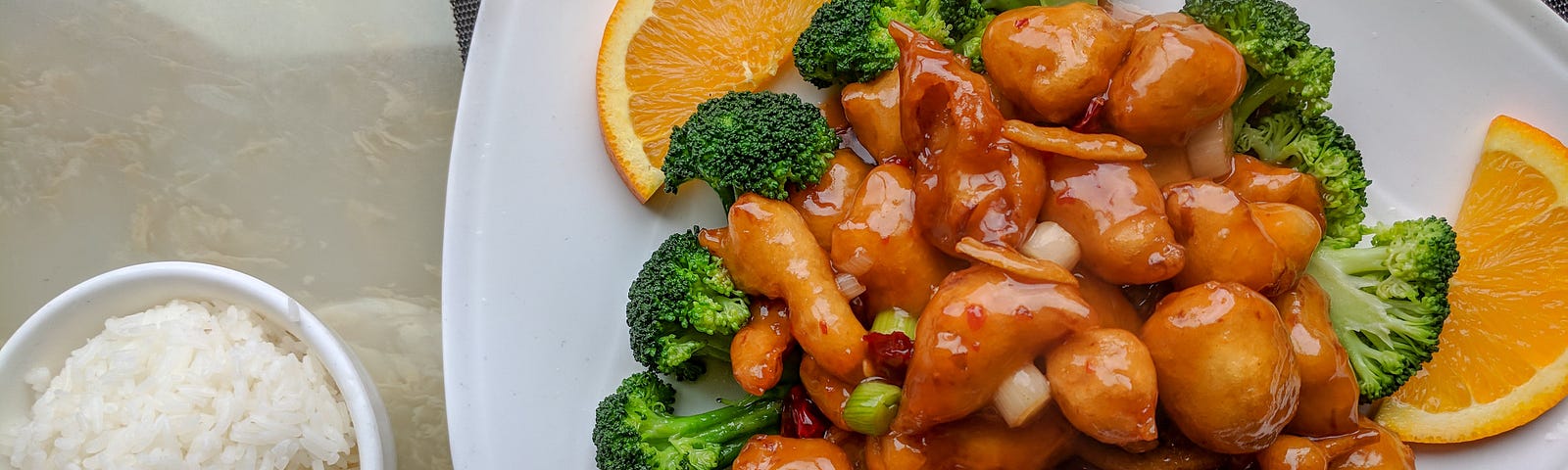 A white fancifully plated with an entree of orange chicken garnished with broccoli and orange slices and a side white bowl of white rice is served next to the plate.