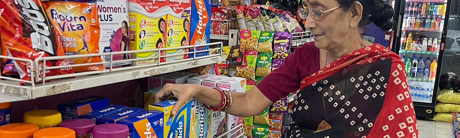 A woman in a tie-and-dye flowing garment is shopping in the Horlicks (processed malt-based powder) aisle at a grocery store. The woman is wearing bangles. There is a strip of masala packages hanging behind her.