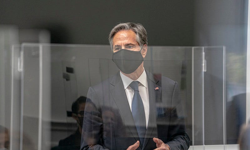 Secretary of State Antony Blinken stands behind a plexiglass barrier, with a face mask, addressing a room of students at the Foreign Service Institute.