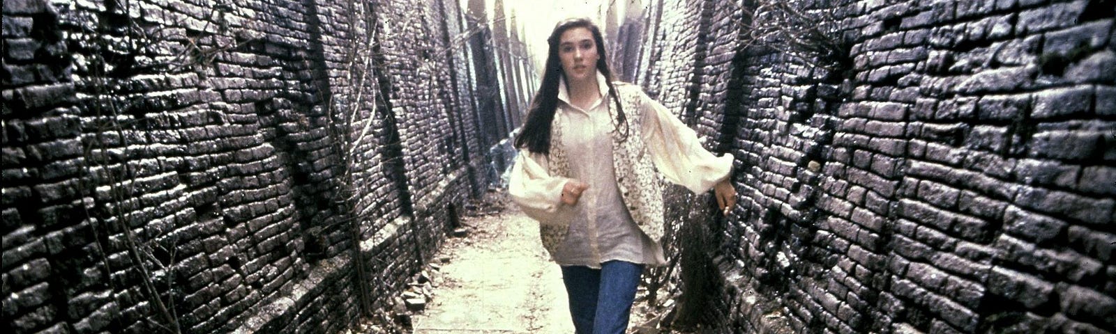A scene from the movie Labyrinth: A young, white woman in a white poet’s blouse, vest, and jeans runs along a desolate labyrinth hallway with broken bricks and dead vines on an overcast day.
