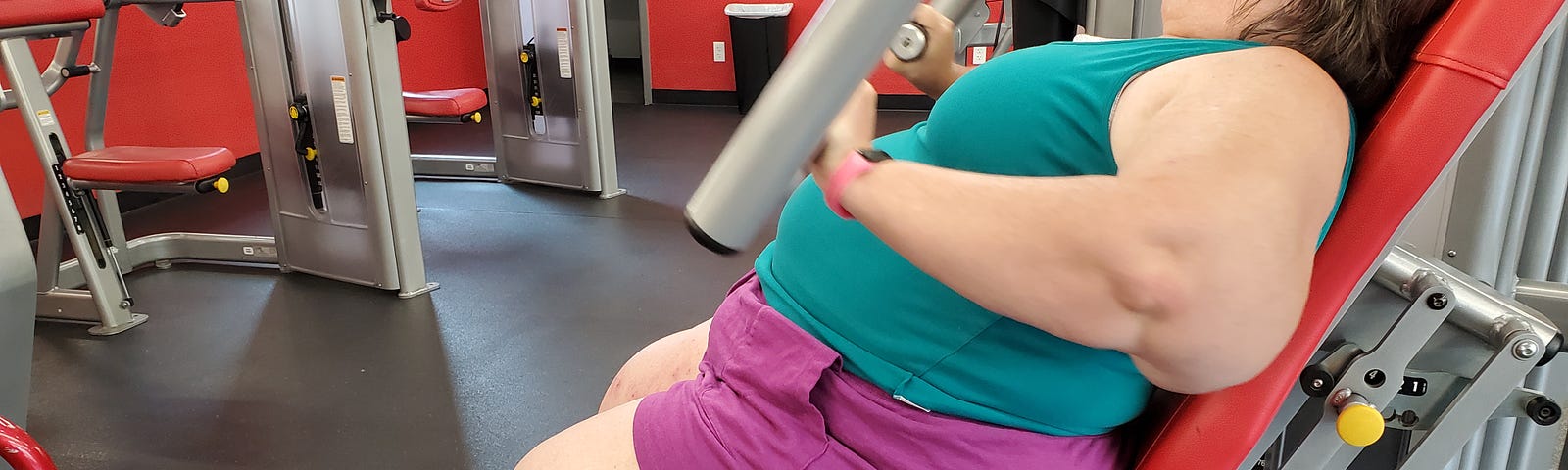 Middle aged fat woman in green tank top and purple shorts using a weight machine in a gym.
