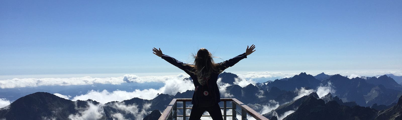 Fearless successful woman standing on bridge joyously stretching arms out,  facing clouds