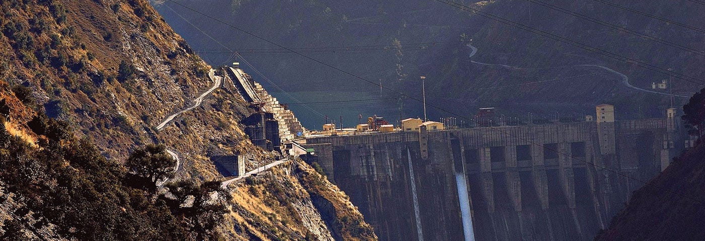 A steep-sided valley with small trees on the hillsides is shown, with sunshine on one slope, and Baglihar Dam mostly in shadow. A white stream of water flows through one of the dam’s sluice gates, and a small amount of the dam’s reservoir is visible in the background. Baglihar is a run-of-river power project on the Chenab River in the southern Doda district of the Indian state of Jammu and Kashmir. This is a closer crop of the photo included in the body text of the article.