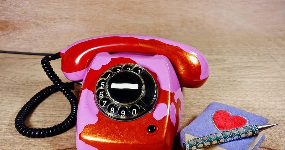 A red and pink rotary phone with a small notepad and bejeweled pen beside it.