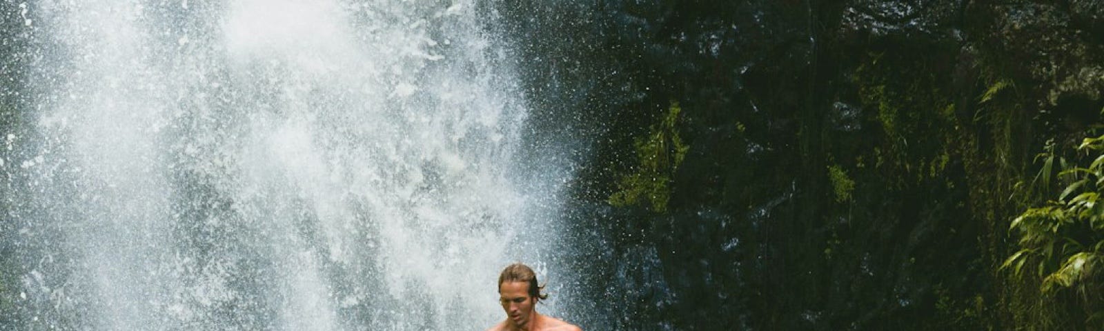 background of half waterfall and half dark green vegetation with a man doing the “dead fish” jump, which makes him look as if he is standing in midair