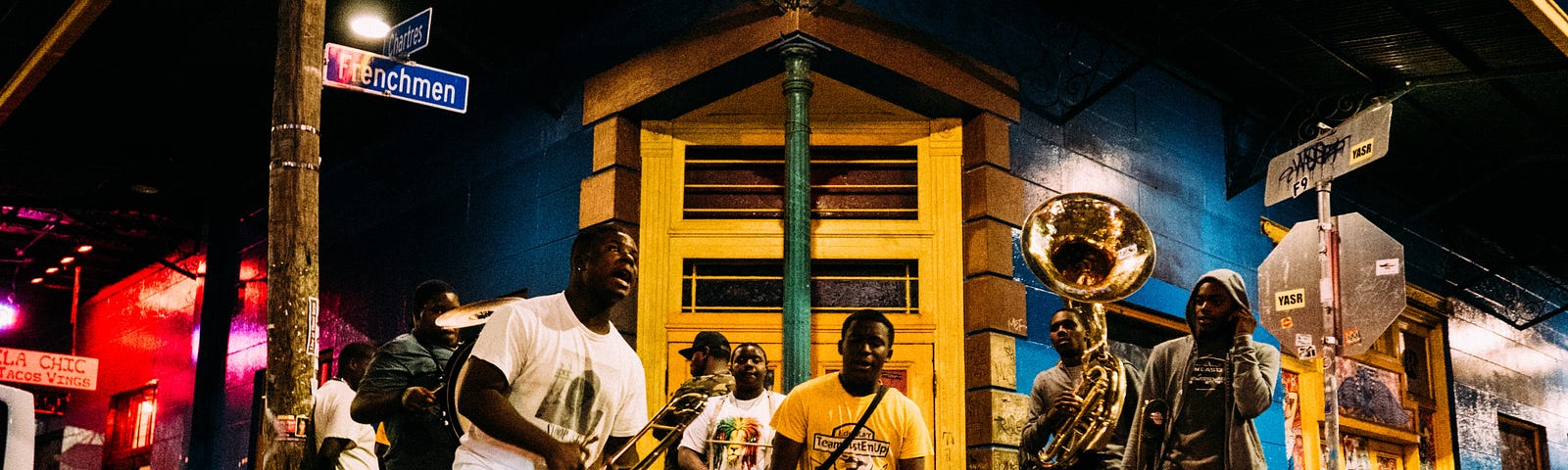 Eight men holding trumpets and standing in front of a shop by the street