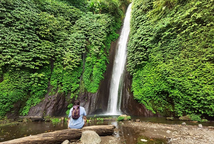 man wearing backpack sitting in front of waterfall surrounded by green vegetation