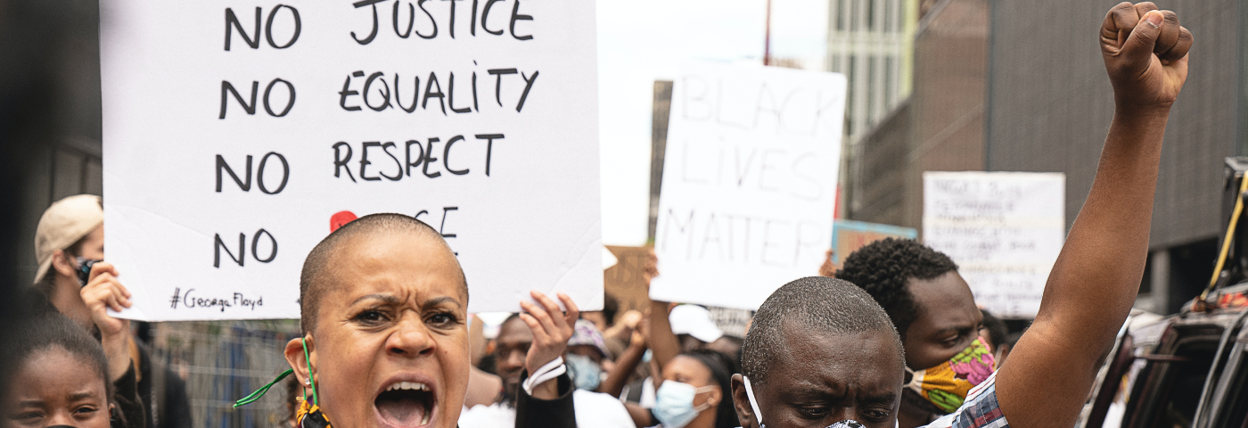 A picture from a protest, a black woman is speaking into a microphone, next to her a black man is raising his fist. Behind them is a poster saying “no justice, no equality, no respect, no peace”