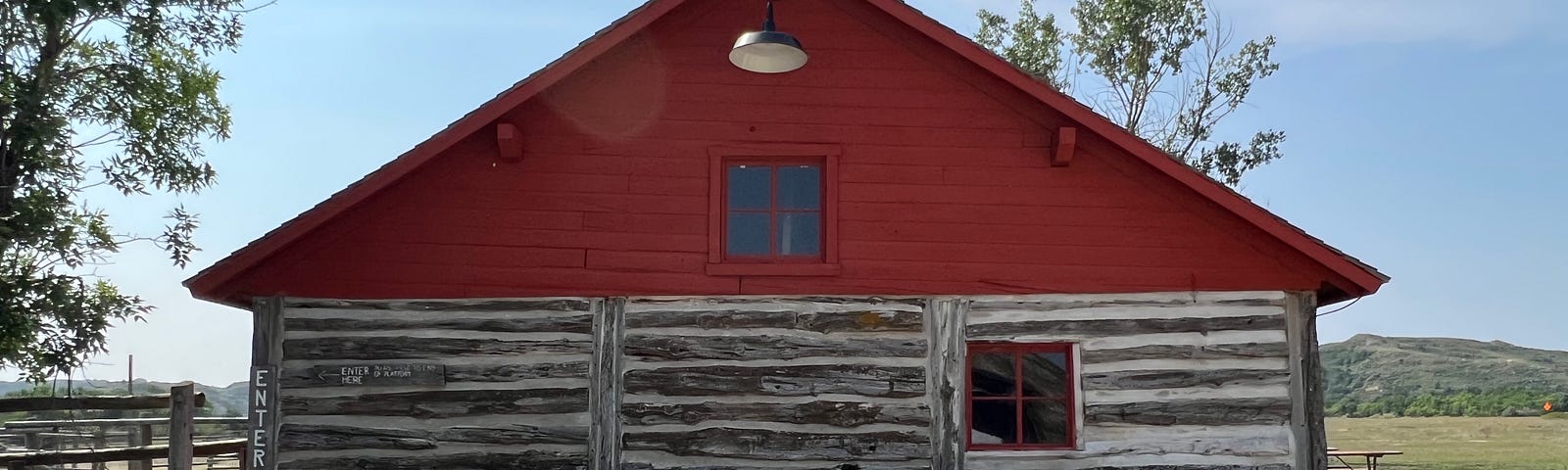 This is a photo of a log cabin with a red roof surrounded by beautiful countryside, green grass, and mountains in the background.