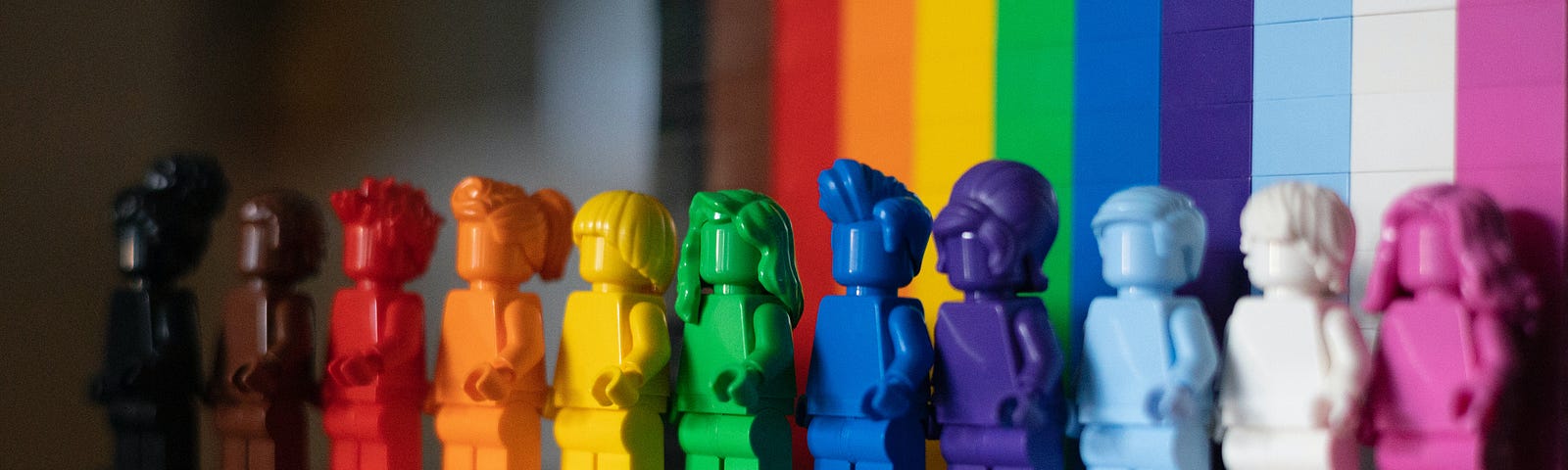 A series of differently coloured lego figurines arranged in the order of the rainbow flag with rainbow lego background