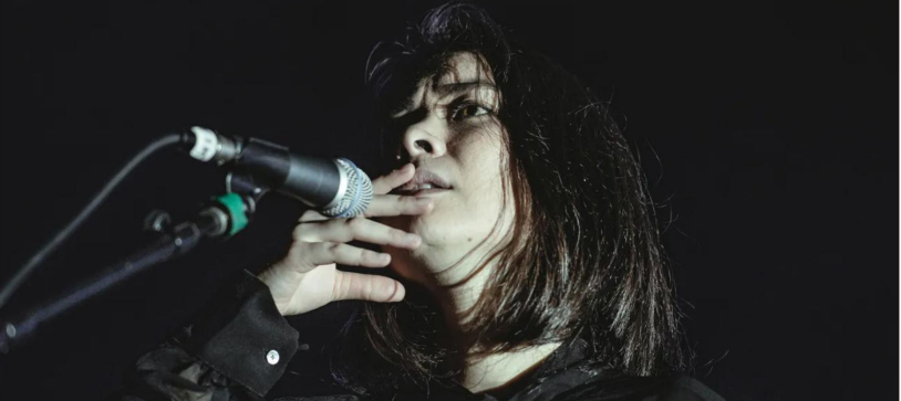 With a black background, a young woman takes up most of the screen. She is holding her lips as she seems to be singing to a microphone that is at the right corner. She has a concerned look but it is unknown what she is looking at.