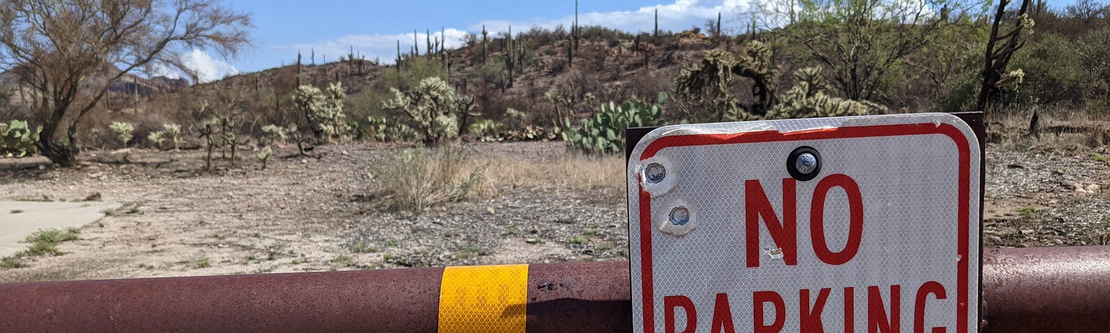 Metal barrier in the desert with sign reading “No Parking Any Time”. The sign has several bullet holes through it.