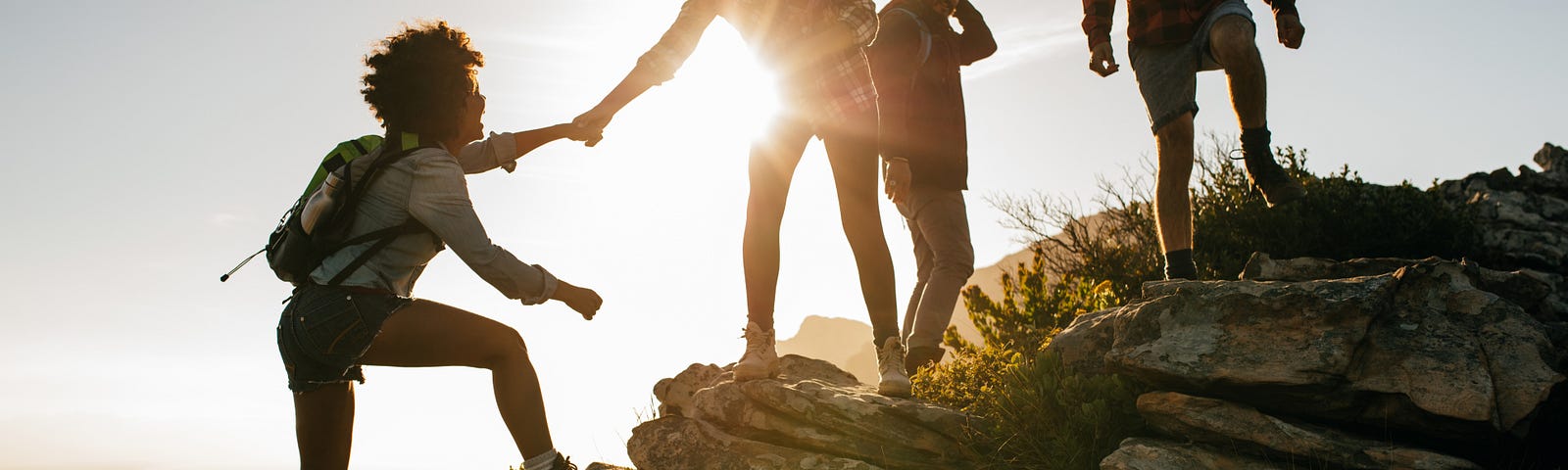 Four adults out hiking together at sunset. Three of them are standing on a low boulder. One of them is holding the hand of the fourth, supporting them as they climb onto the boulder to join the others. All of them are looking happy to be there.
