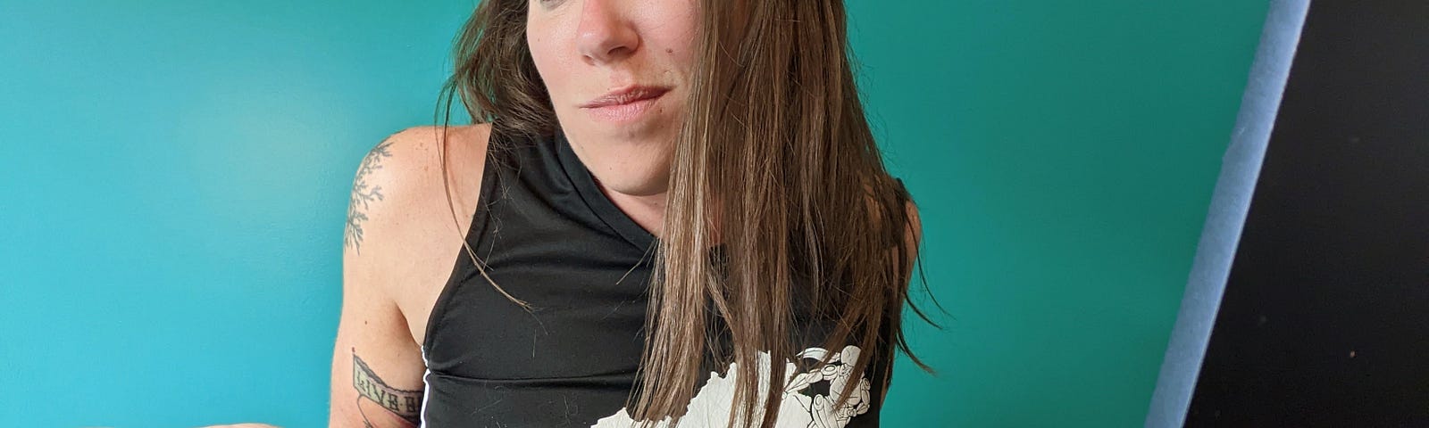 Heather Gioia, white female with long brown hair, is sitting in front of a green wall with a laptop. Heather is shrugging her shoulders. She is wearing a black top with the Virginia All Stars Roller Derby logo on the front in white.