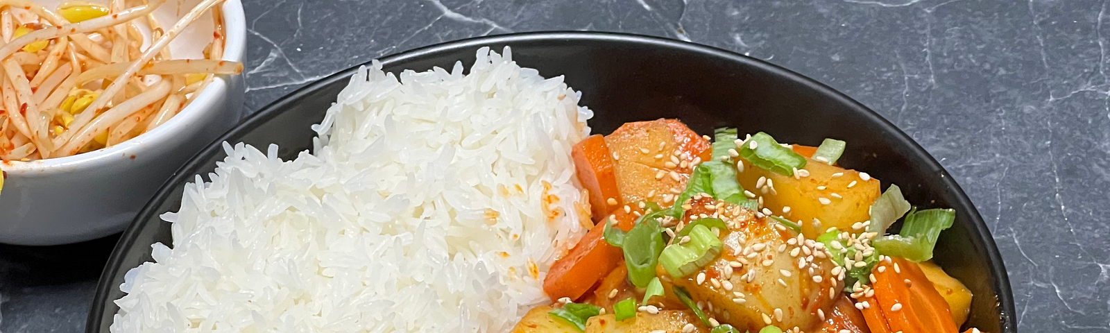 Korean chicken and potato stew served with white rice, spicy bean sprouts and topped with green onion on a black marble countertop