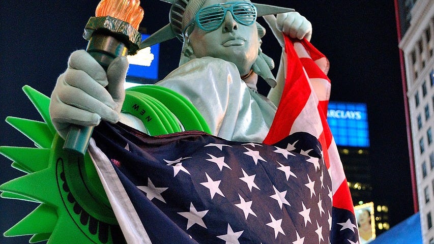 In Times Square, a mock Statue of Liberty is attired in commercial items, such as sunglasses, a flag, and clickbait-y things.