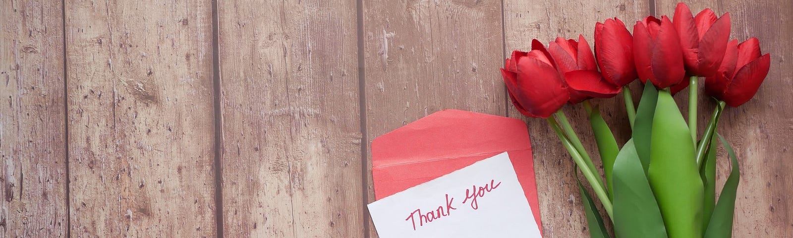 Against a background of wooden boards, a twine-wrapped bundle of red tulips sits beside a small hand-written note that reads “Thank you,” paired with a red envelope.