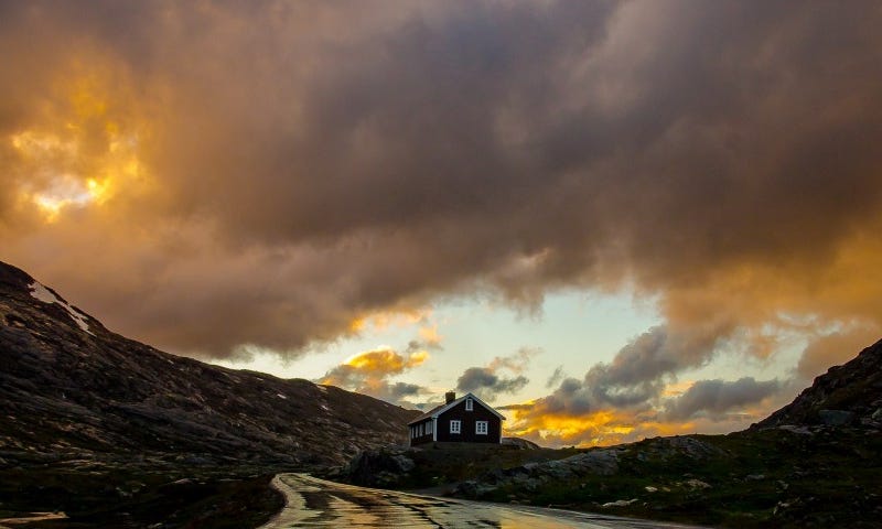 A rain-soaked road leading to a wooden hose on a hill surrounded by mountains and a cloudy sky.