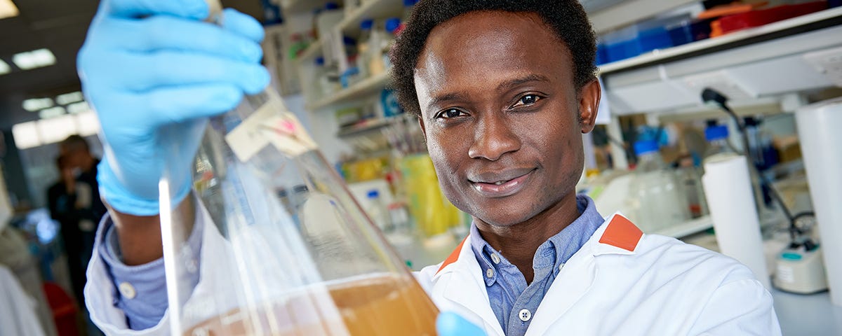 Dr Oluwapelumi Adeyemi wearing a white lab coat and blue gloves holding a beaker with liquid in it