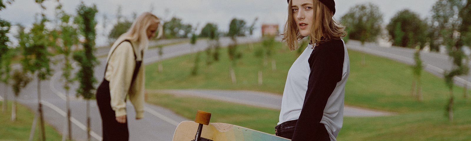 woman with shoulder-length hair holding a rainbow skateboard. another woman skates near her.