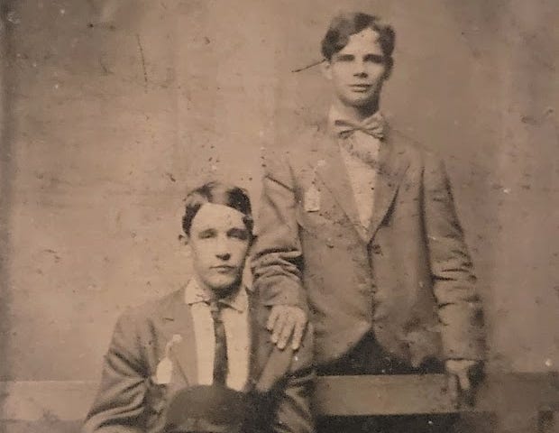 a posed tintype photograph of two young men wearing suits one is seated on a bench holding a hat while the one is standing behind with his hand resting on the others shoulder both have serious expressions on their faces