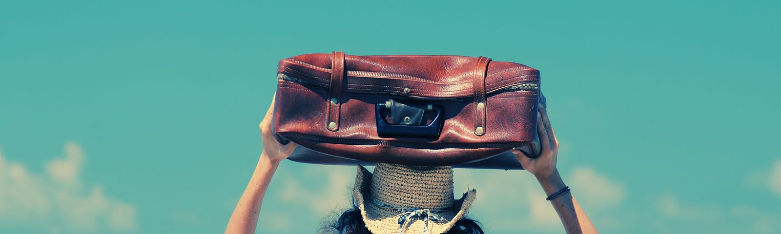 Woman with suitcase on her head overlooking the ocean and bright blue sky.