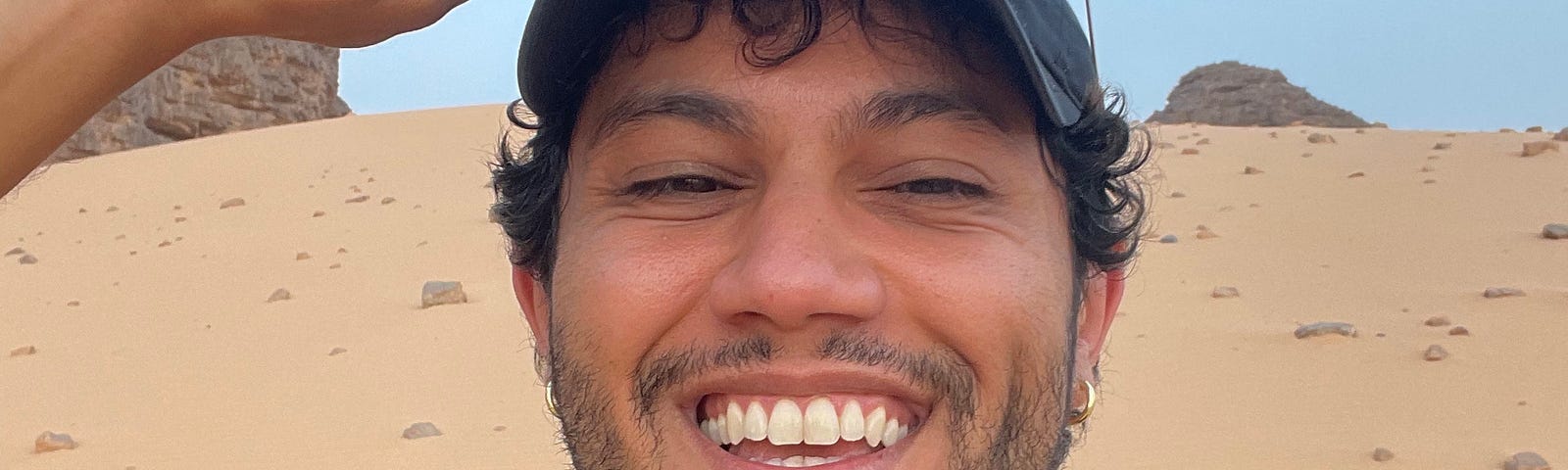 The image features an Amazigh person with brown skin, dark curly hair, and a trimmed beard, standing outdoors in a desert setting. They are wearing a navy-blue t-shirt and a black baseball cap. The person holds their sunglasses, which are black with thick frames, on top of their head with their right hand while smiling broadly, showing their teeth. Their eyes are slightly squinted, and they look directly at the camera, radiating warmth and happiness.