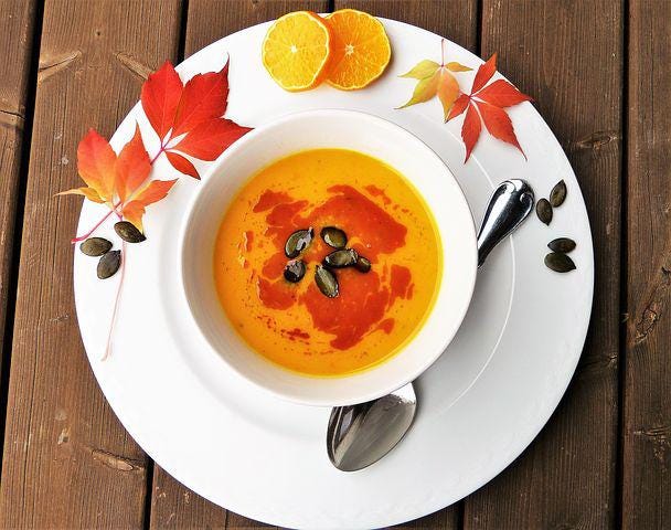 A white dinner plate, with autumn leaves and a slice each of orange and lemon on the side, and resting on the plate is a white bowl full of reddish / orange soup.
