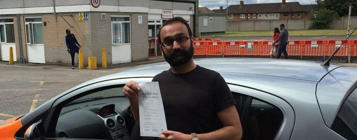 Brown man with beard and glasses in black clothes standing in front of a a learner car holding a piece of paper.